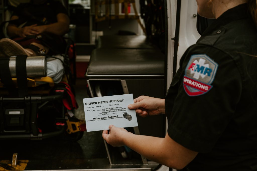 A paramedic stands at the back of an ambulance and reads a blue card that states Driver Needs Support