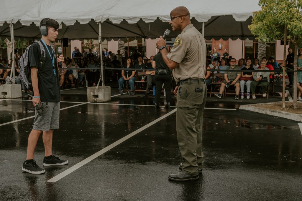 A police officer holding a microphone talks to a young client wearing headphones in front of an audience sitting under a tent. The ground is wet with rain