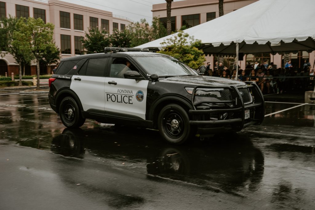 A police vehicle is parked in front of an audience sitting under a tent. The ground is wet with rain