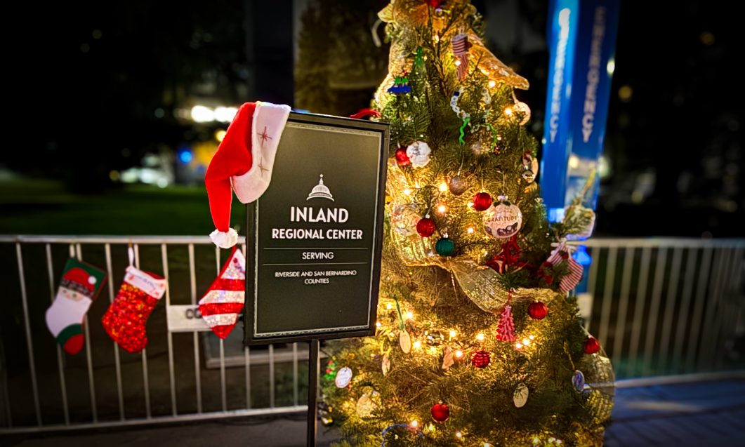 Christmas tree adorned with lights and ornaments beside Inland Regional Center sign, Santa hat on top.