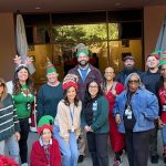 Group of staff and community members wearing festive holiday hats and accessories while posing together outdoors in front of a building entrance.