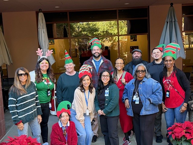 Group of staff and community members wearing festive holiday hats and accessories while posing together outdoors in front of a building entrance.