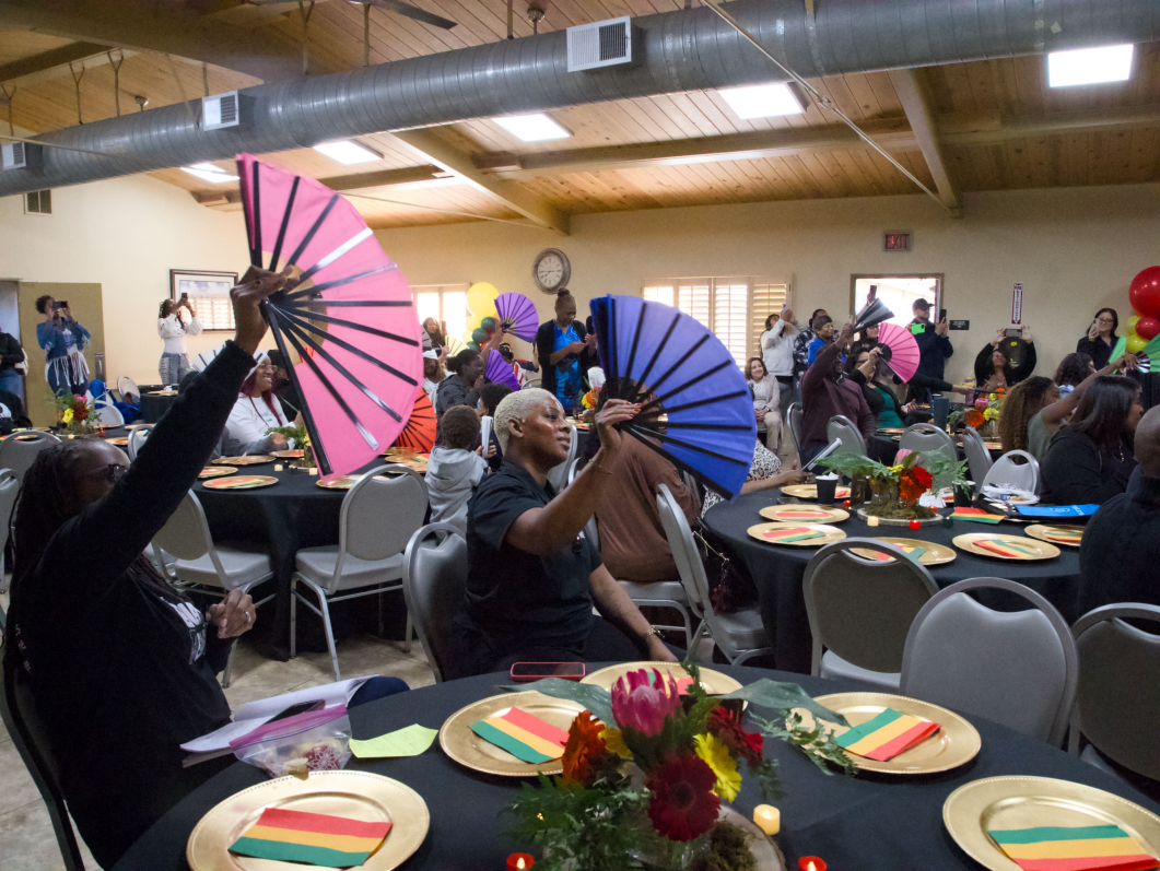 Attendees seated at decorated banquet tables waving colorful folding fans during a lively indoor community gathering or celebration.