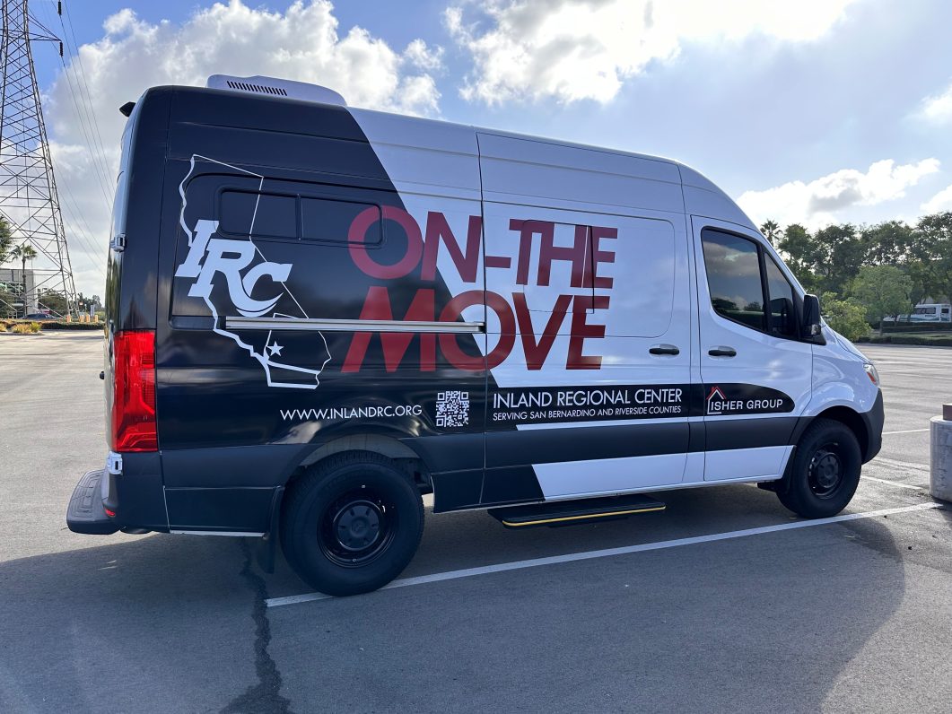 A picture of the the right side of a black and white Sprinter cargo van displaying the classic IRC Inland Regional Center logo along with the words ON-THE-MOVE, links, and a QR code in a parking lot.