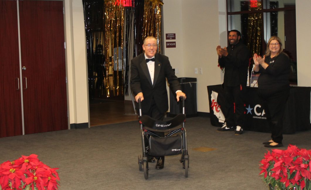 A person in a tuxedo walks into a room decorated in gold and black streamers using a four wheel walker. Two people in the background are cheering them on