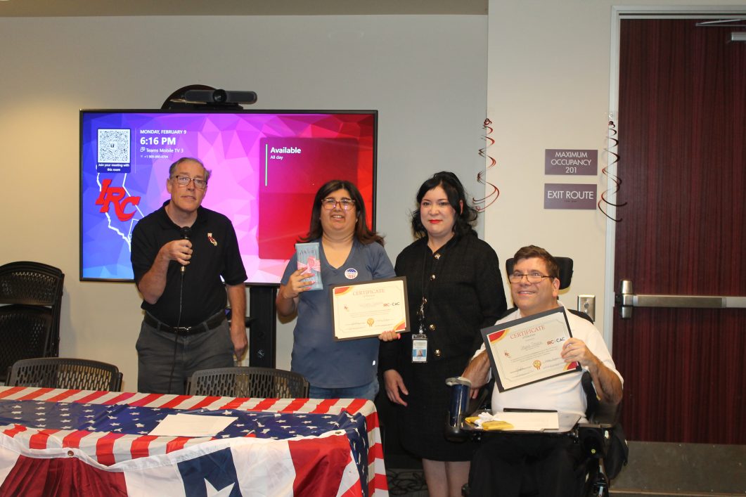 Four people posing during a certificate presentation at a community recognition event. Two participants hold certificates while standing beside a person speaking into a microphone and another participant using a powered wheelchair. Patriotic table decorations and a presentation screen are visible in the conference room background.