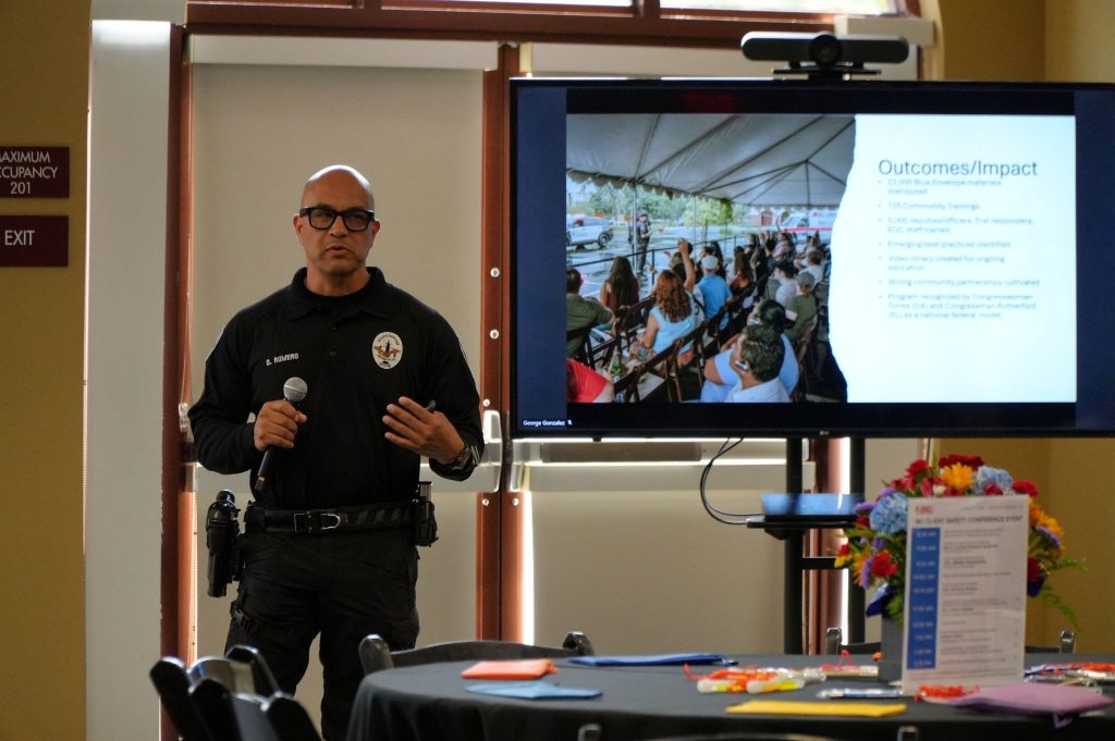 A police officer holds a microphone in a conference center and stands near a large television that is displaying information
