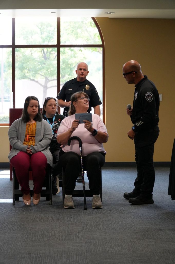 Two police officers stand near three sitting audience members. One person holds a blue envelope while speaking to an officer