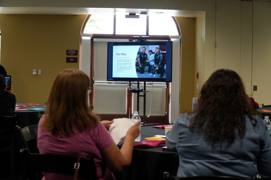 Two people sit at a table and look towards a large television that is displaying a presentation.