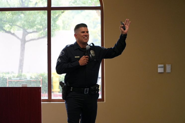 A smiling police officer speaks into a microphone in one hand and holds an object up high in his other hand. There is a large window behind them