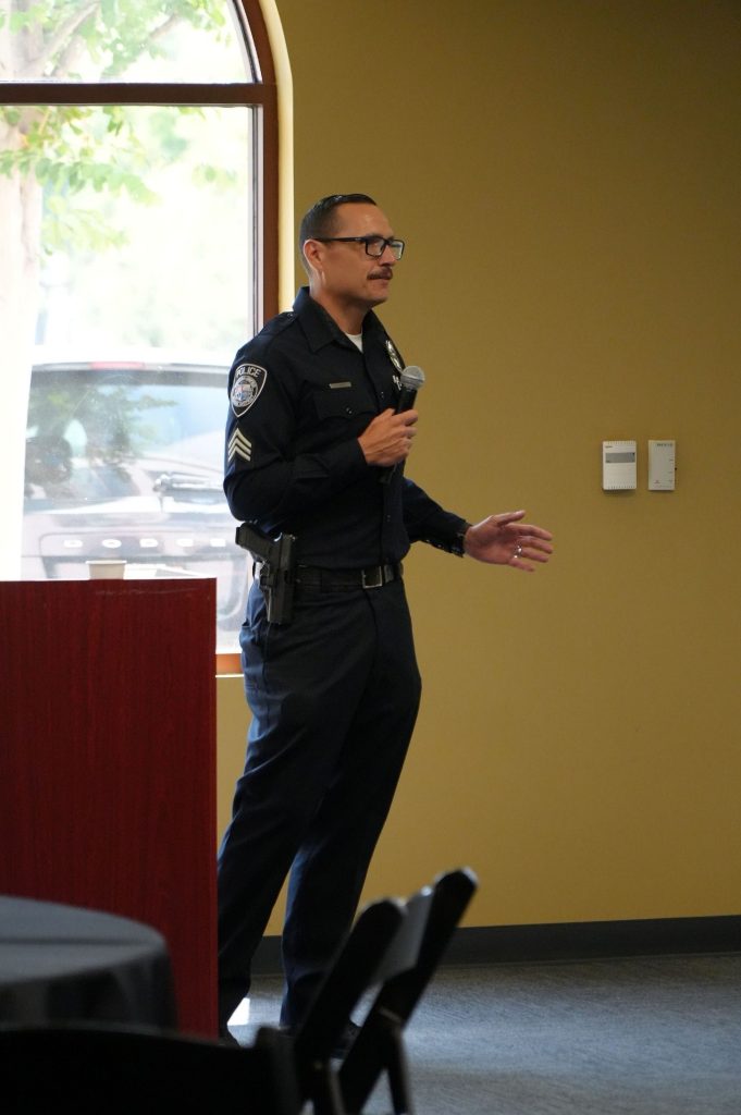 A police officer with glasses holds a microphone in one hand and motions with their other hand