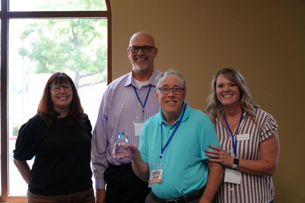 Four people pose for a photo near a large window. Two people wear lanyards and one person holds an award