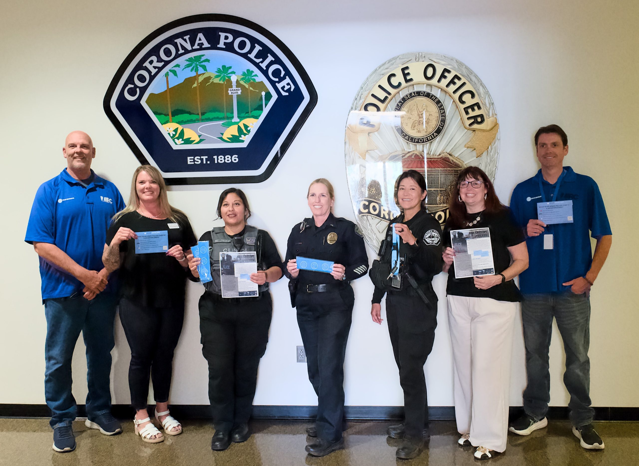 Seven people pose for a photo in front of Corona Police Department signage. Three people are dressed in police uniforms and several people are holding Blue Envelope promotional materials.