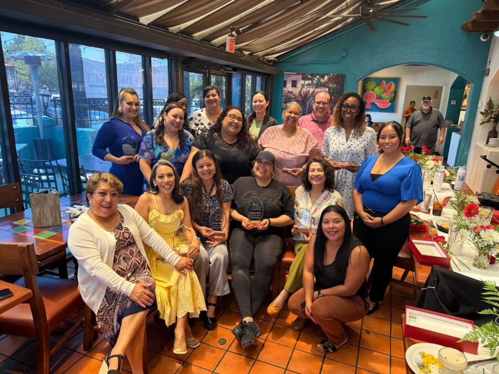 A large group of people post for a photo inside of a restaurant. Several people hold glass trophies and flower arrangements sit on top of the tables