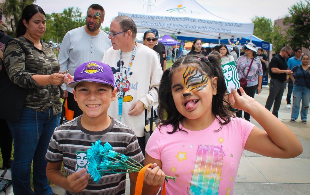 Two children pose for a photo at an outdoor resource fair holding Starbucks gift cards. One is wearing a purple hat and striped shirt and the other has tiger face paint and a pink shirt. Several other people are in the background
