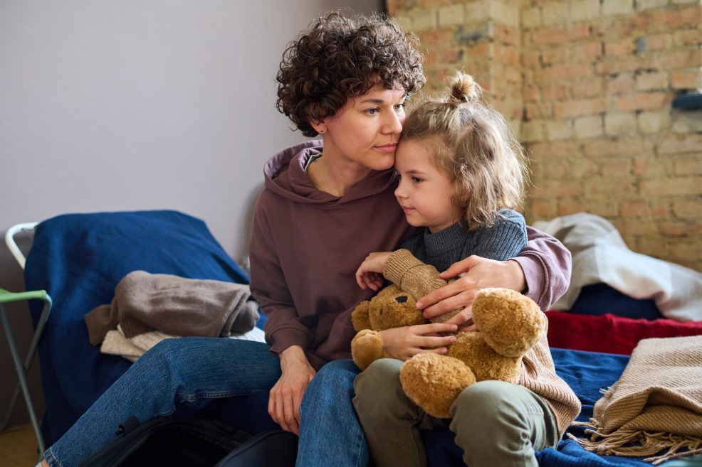 Young mother giving hug to her child with brown soft teddy bear while sitting on a cot