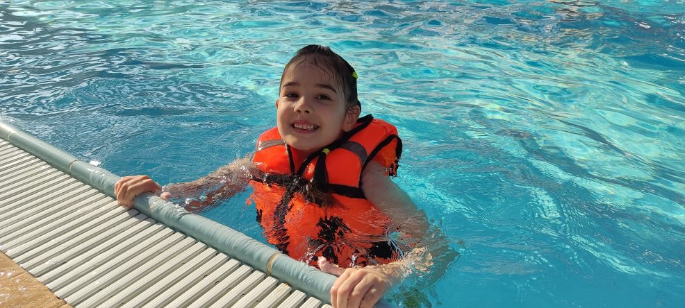 Child wearing life jacket smiling in pool on sunny day.